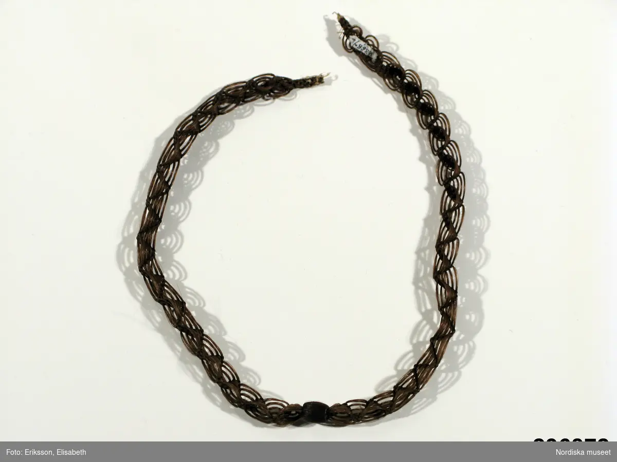 Kedja i glest hårarbete med överklädd kula på mitten,  är ej monterat med metallbeslag och funktionen framgår inte, möjligen halsband eller klockkedja.
/Berit Eldvik 2009-12-01