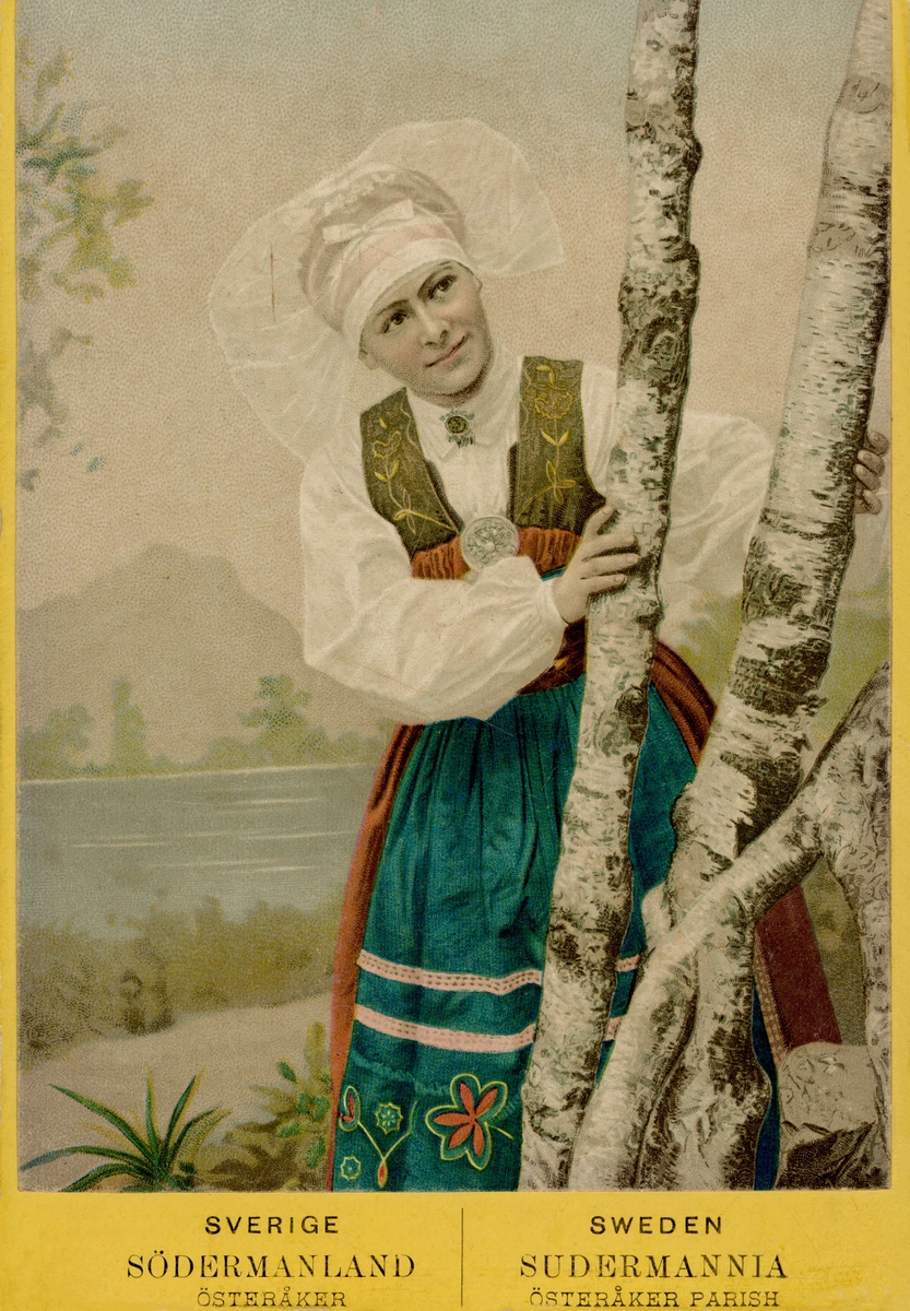 Kolorerat porträttfotografi av som kvinna tittar fram bakom björkstammar. Hon är klädd i folkdräkt från Österåker i Södermanland. Dräkten sydd som en nationalromantisk rekonstruktion av originalplagg kring 1900.