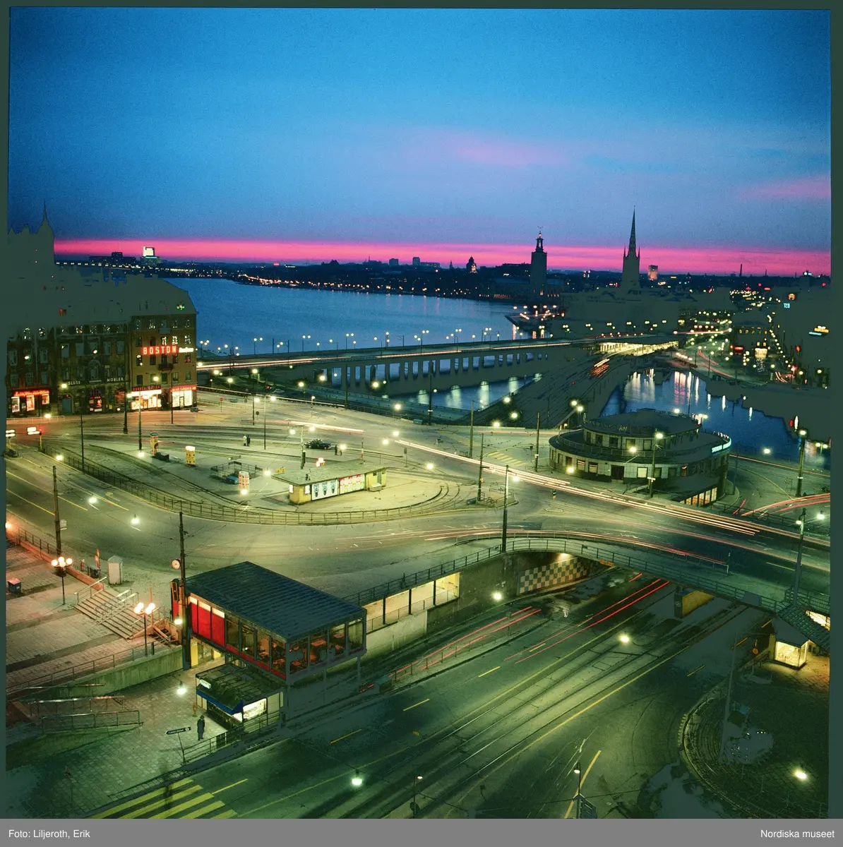 Slussen, Stockholm. Stadsbild med vy &ouml;ver Slussen, i bakgrunden Riddarholmen med kyrkan och Kungsholmen med Stadshuset i f&ouml;rgrunden. Det &auml;r kv&auml;ll, m&ouml;rkret har s&auml;nkt sig och gatubelysningen t&auml;nts, men himlen i v&auml;ster &auml;r fortfrarande orange och rosa efter solnedg&aring;ngen.
