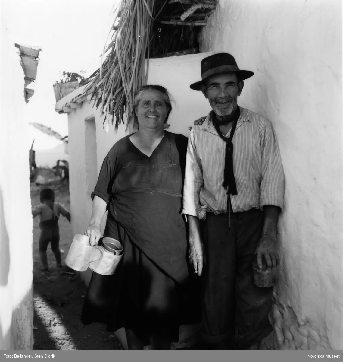 Fiskeby utanför Torremolinos, Spanien. Ett par i övre medelåldern står i en gränd och skrattar mot kameran. Deras kläder i slitna och de håller krus och kärl i händerna. Mannen håller en cigarett mellan fingrarna. I bakgrunden skymtar en liten pojke utan byxor.