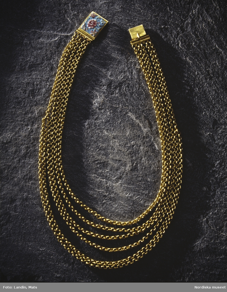 Halskedja av guld med lås i emalj. Låsets baksida försett med Kung Karl XI:s namnschiffer. Föremål ur Nordiska museets samlingar invnr:
219188.
