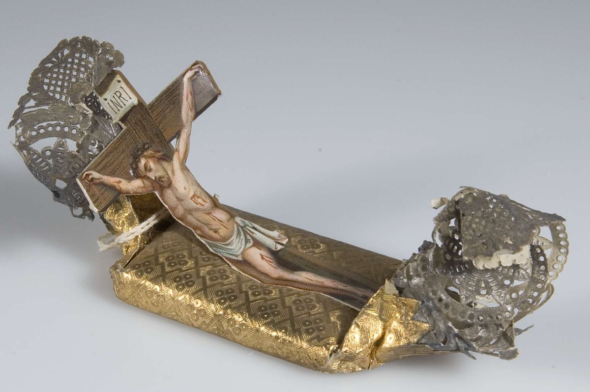 Begravningskaramell, omslag av mönstrat guldpapper och genombrutet silverpapper. Märke i färg med Jesus på korset. 