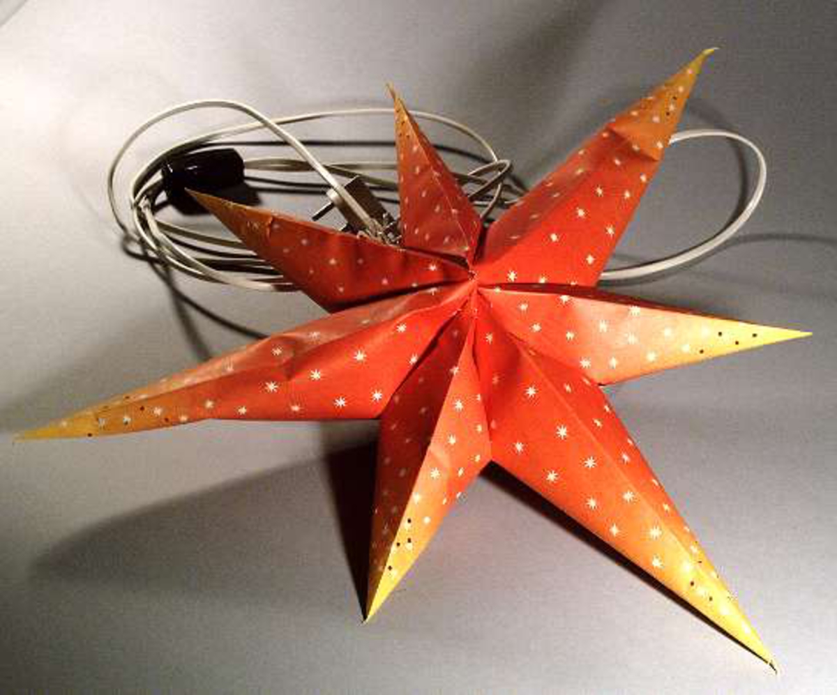 Adventsstjärna av papper med elektrisk lampa och sladd. Stjärnan rödorange med vita stjärnor och perforerad i ändarna.
