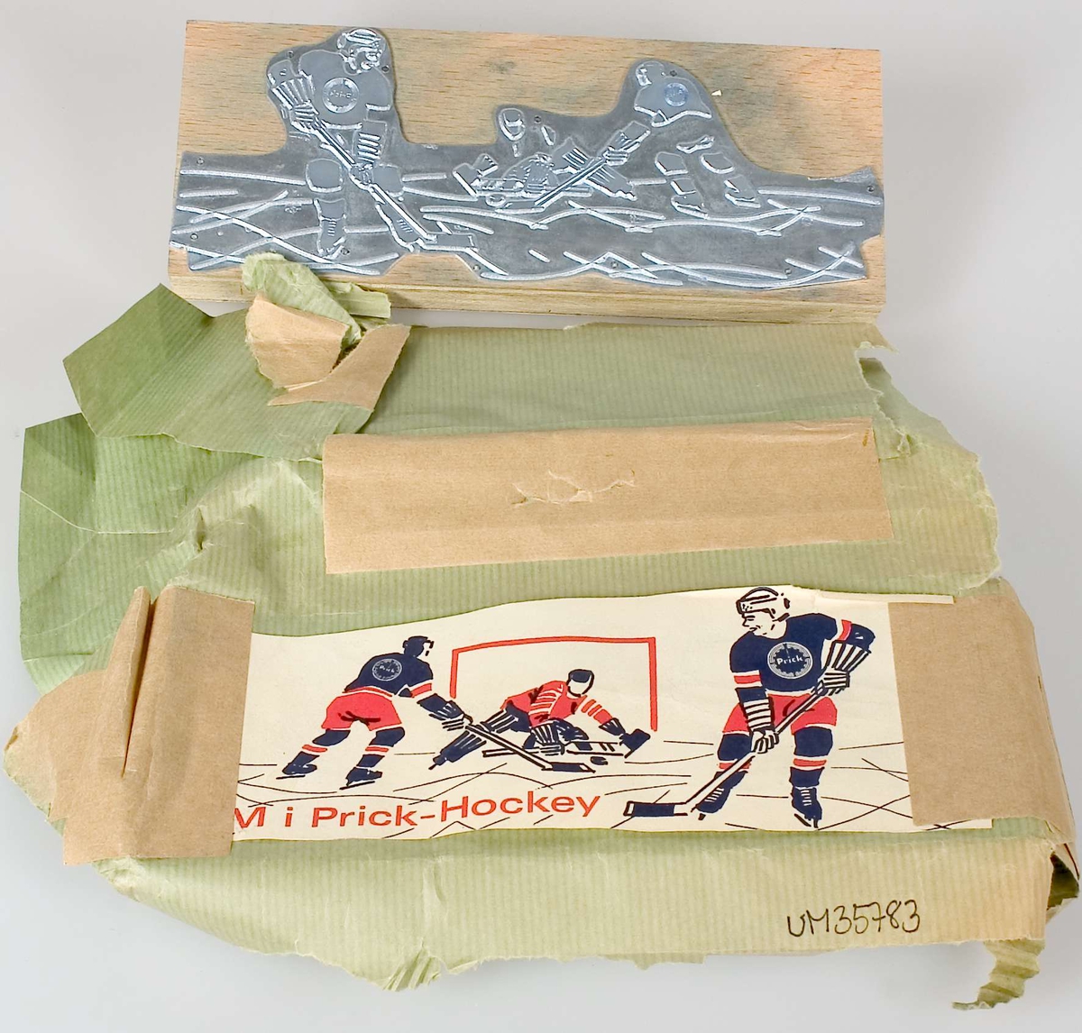 En kliché av metall, monterad på en platta av skiktlimmat trä. Har varit förpackad i ett  grönt omslagsapper på vilket ett exemplar av den tryckta produkten är fäst med tejp. Tryck i rött och blått, tre ishockeyspelare samt texten: VM i Prick-Hockey. Logotypen Prick på två av ishockeyspelarnas tröjor.