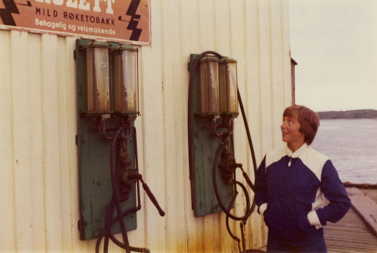 En kvinne ser på en reklameskilt for Rulett tobakk som sitter ovenfor to bensinpumper.