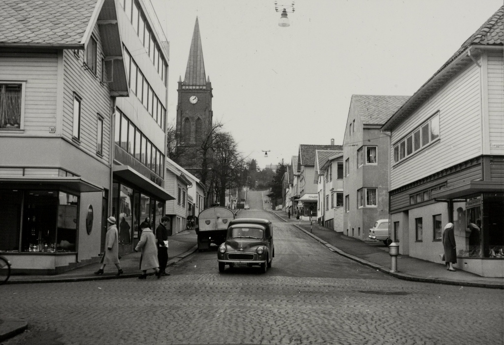 Motiv fra krysset mellom Kirkegata og Langgata i Sandnes sentrum våren 1960 med Sandnes kirke i bakgrunnen. Hjørnet til venstre rommer gullsmed Christophersen og til høyre ligger skomaker Vagle. Nybygget bortenfor hjørnehuset tilhører Christophersen. Bilen i krysset er en Morris Minor varebil.
