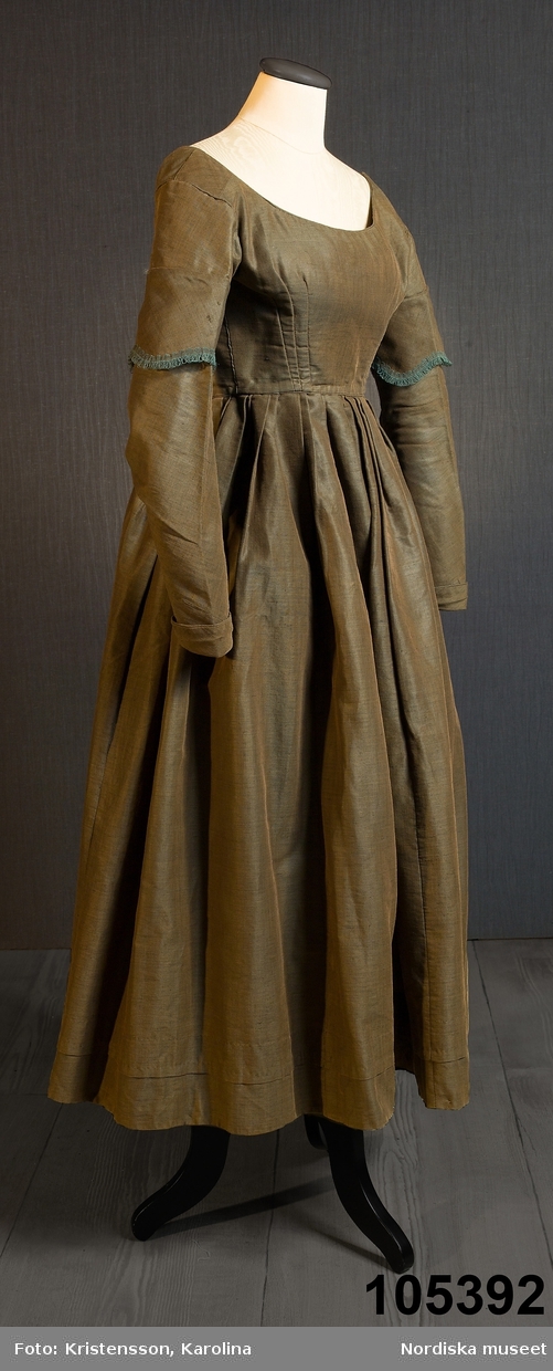 Hel klänning i 1830-talets modesnitt, av fint halvylletyg i kypert med varp av ljusbrunt bomullsgarn och inslag av grönt tunt glansigt redgarn som ger en grönskimrande yta. Livet går ned nästan till midjehöjd, figursytt med 2 framstycken med bakåtflyttade sidsömmar, ett ryggstycke med 6 inprovningar fördelade i två grupper. Vid båtringad halsringning, knäppning med tätt sittande hakar och hyskor. Isydda långa ärmar svängda med 2 sömmar  med 3 veck vid armbågen. Ärmsprund knäppt med hyska och hake samt 4 cm uppslag med rundade hörn. Upptill på ärmen besättning av tyget med stora rundade uddar kantade med klargrön bomullsfrans.  Passepoaler av tyget i alla sömmar. Livet fodrat med skotskrutig bomullslärft i rött, grönt, gult och vitt och i ärmarna ett smalrandigt bomullstyg i blått och rosa. Kjolen sydd av  5 våder, lagda veck runtom i midjan. sprund mitt fram. Uppsytt veck  en decimeter från fållen. Skoning med brun bomullslärft. Handsydd.
Har burits 1838 som ungmorsklänning, alltså av bruden dagen efter bröllopet, av Maria Margareta Mårtensdotter från Kysings, Vall sn på Gotland. Möjligen tillsammans med sidenhalsklädet 105385.
Anm. I  gott skick.
OBS Klänningen sitter bakochfram på fotona, knäppningen är fram.
/Berit Eldvik 2011-06-13