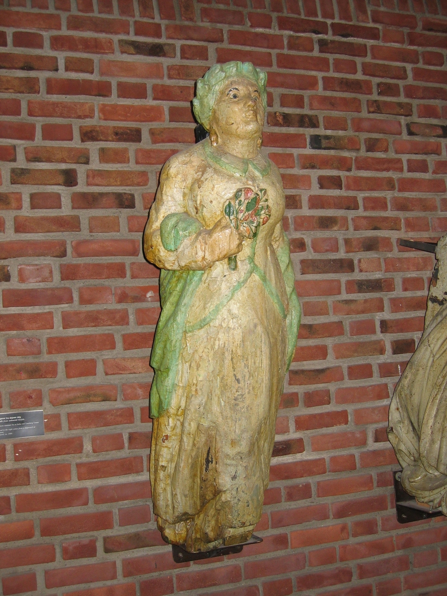 En klassisk gallionsfigur, venstre arm ned langs siden, høyre arm bøyd med en blomsterbukett i hånden mot brystet. Sort hår, hvit kjole med grønn kant, blomster i høyre hånd mot brystet.
