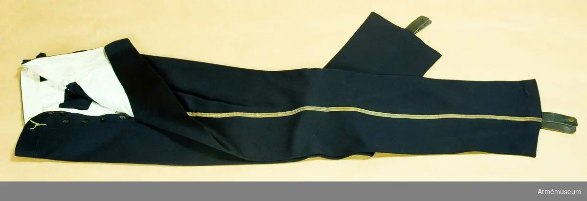 Mörkblått kläde, guldgalon m/1895 längs yttersömmarna, två sidofickor. Svarta läderhällor att knäppas under foten med mässingknappar. Foder av vitt siden.