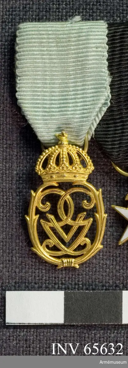 Grupp M II.
Jubileumsminnestecken, krönt monogram i guld. Ljusblått medaljband.