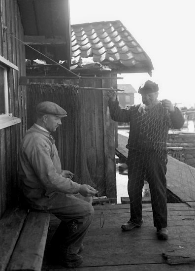 1924. Två fiskare med garn, på bryggan vid sjöbodar.