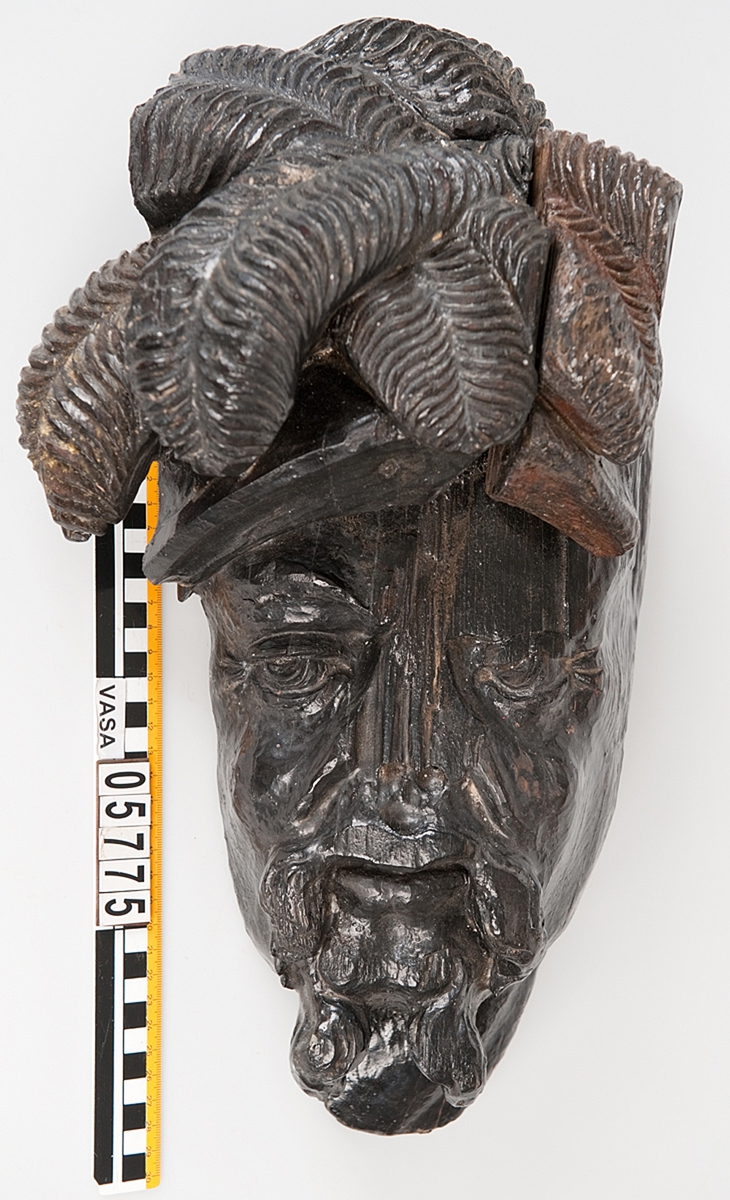 Del av skulptur. 
Huvudet till en högrest krigare med stor mustasch. På huvudet sitter hjälm med långa vajande plymer.