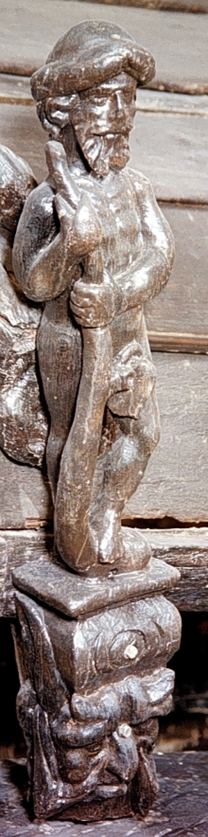 Skulptur av en halvnaken man, så kallad vildman, stående på en hög sockel.

Mannen har krusigt skägg, stora mustascher och långt hår. På huvudet sitter en tjock lagerkrans. Skulpturen är frontalvänd. Utmed höger sida håller mannen med bägge händer en stor klubba på vars böjda nederända han stöder vänster fot, så att kroppstyngden vilar på höger ben. Från klubban utgår en kvist med vilken den nakna mannen nödtorftigt skyler sig.



Figuren är rundskulpterad bortsett från bakhuvudet som är något avplanat. Mannen står på en rektangulär och horisontell platta som bildar övre delen av en kraftig sockel. Sockeln är till största delen formad som en grotesk maskaron med stora ögon, bred näsa och spetsiga åsneöron. Mellan ståplattan och maskaronen ses en kraftig vulst prydd med koncentriska skåror. Nedtill är sockeln snedskuren. Baksida är plan med nedtill ett på höjden rektangulärt urtag och har utgjort anliggningsyta. Basen är horisontal.



Skulpturen är mycket välbevarad.



Text in English: Sculpture of a half-naked man on a high pedestal, shown en face.

He has a curly beard, large moustache, and wears a thick laurel wreath on his head, he has long hair. He holds a large club in both hands at his right side, the club''s lower end is curved, and he rests his left foot upon it, while his right supports the weight of his body. A sprig from the club enables him to partially conceal his nakedness.



The figure is sculpted in the round except for the back of the head which is rather flat. The man stands on a rectangular horizontal entablement. The pedestal mainly comprises a grotesque mask with large eyes, a broad nose, and donkey-like ears. Between the entablement and the mask there is a beading decorated with concentric grooves. The pedestal mask was carved obliquely. The back is flat.



The sculpture is very well preserved.