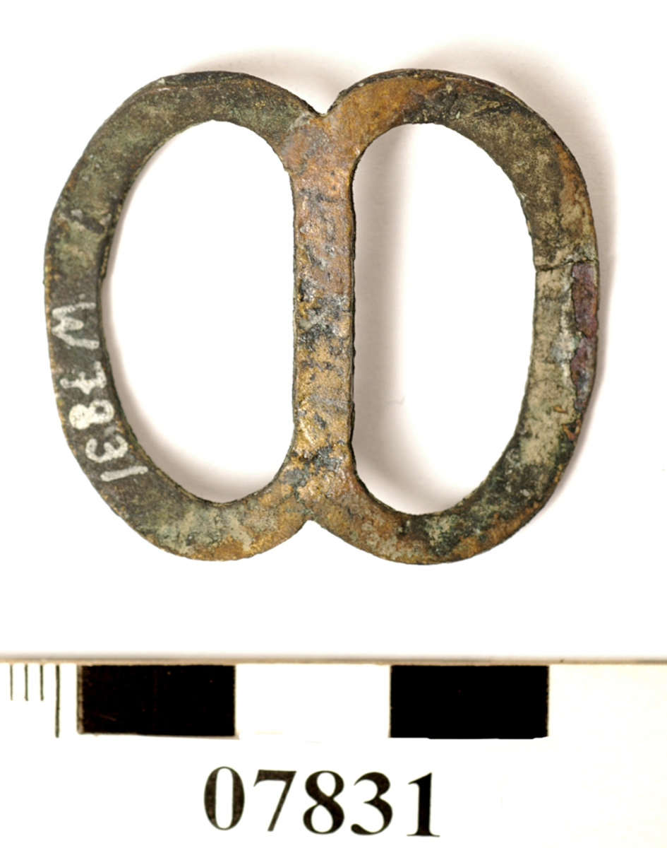 Sölja, spänne. Formad som en åtta. Lätt böjd på mitten.

Text in English: One metal buckle.
One side of metal (probably front) is convex, the other side (probably back) is flat, with defined edges.
The whole piece is concave, the center bar sits lower than the sides. When viewed from the side, it resembles a wide V. When viewed from the front or the back, it resembles a flattened figure 8.
Handmade out of what appears to be one piece of metal, two solder points visible.