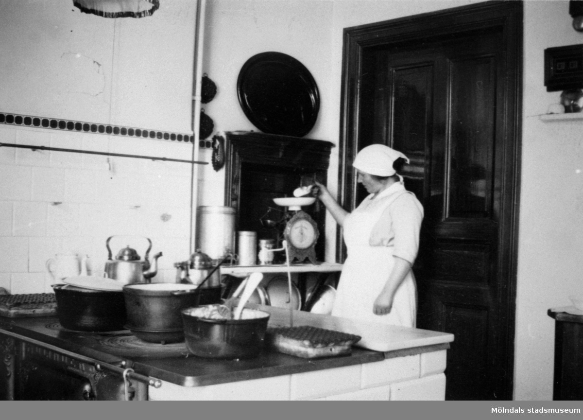 Hilma Olsson arbetar i köket och mäter upp något på en våg i köket i Stora Skolan på Stretereds skolhem okänt årtal. Hennes son (Åke Olsson, född i början av 1920-talet) var fosterson i Vommedal.