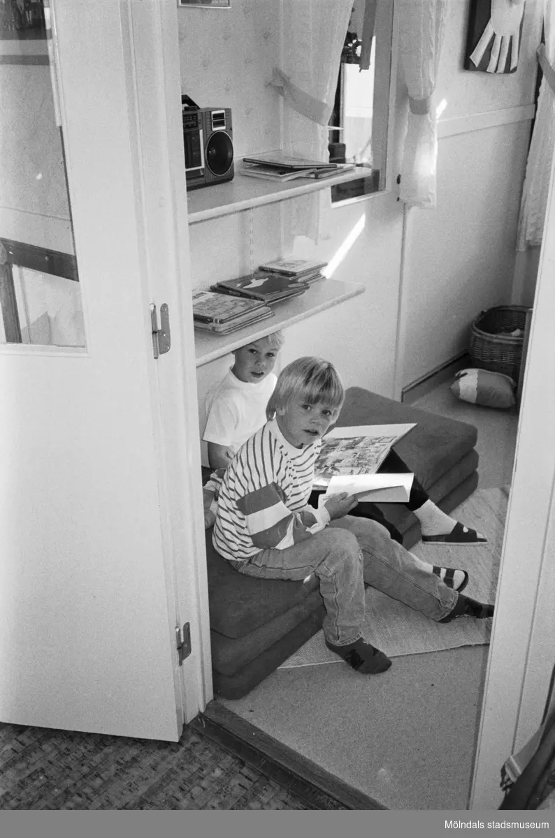 Två pojkar sitter på tre mindre madrasser i ett rum och bläddrar i var sin bok. De är fotograferade ifrån ett intilliggande rum, genom en dörr-öppning, och de sitter precis innanför. Katrinebergs daghem, 1992.