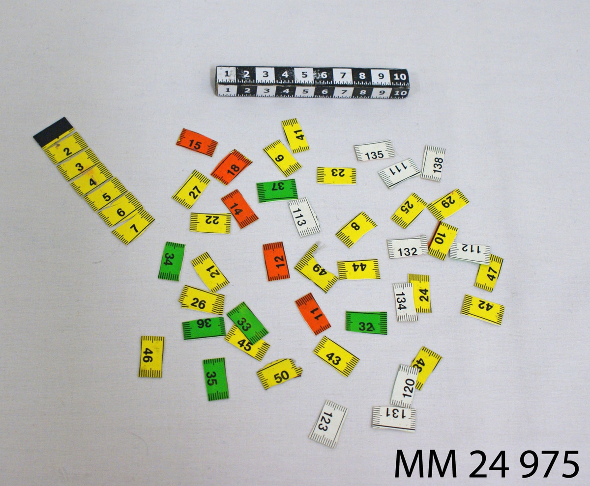 Måttband av plast, sönderklippt i 50 stycken centimeterstora delar. Änddelens kant är försedd med metallplatta. Ena sidan av måttbandet är vit med svart markering, andra sidan är gul, grön eller orange med svart markering.