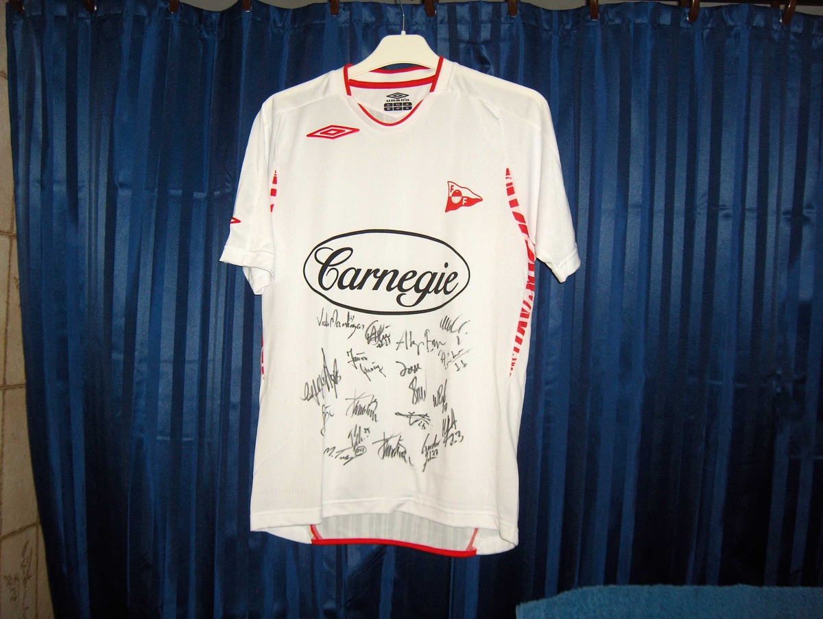 Hvit fotballtrøye med røde kanter med signering av FFK-spillerne på front nedre del under sponsorlogoen til Carnegie.