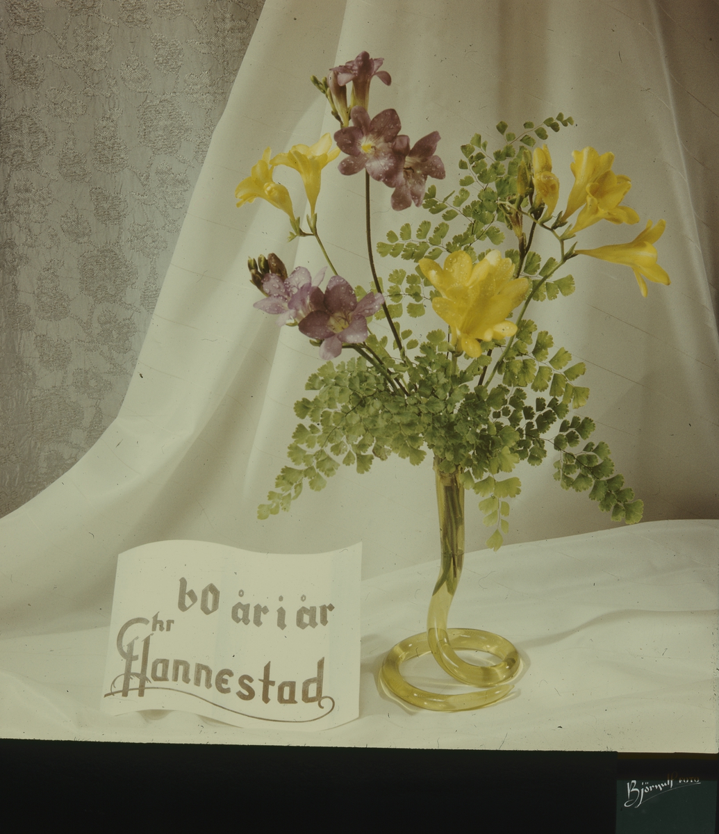 Kinoreklame fra 1962 med blomsteroppsats av fresia i gullforgylt vase, skilt med påskriften "60 år i år" og Chr. Hannestad sin logo, med hvite draperier i bakgrunnen.