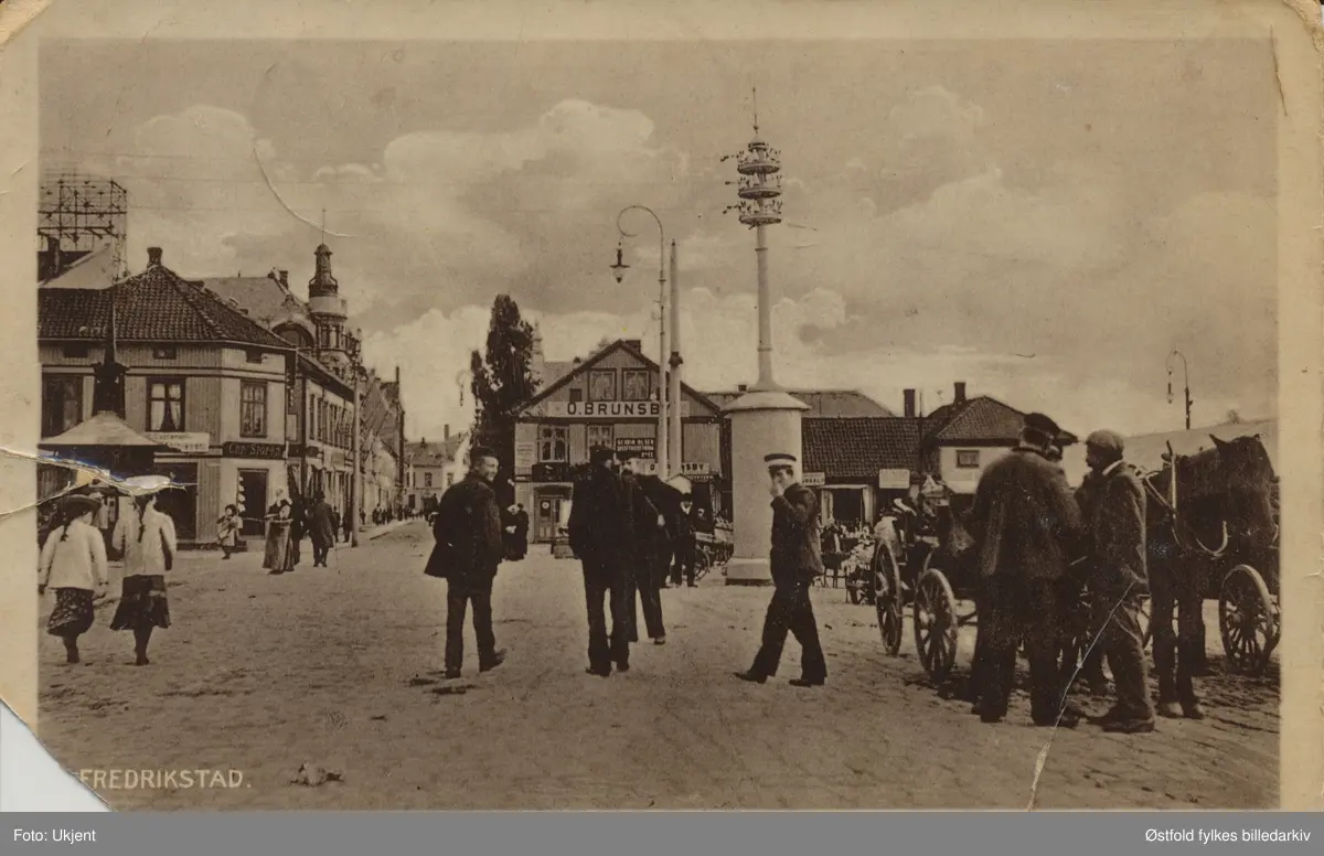 Torvet. på Vestsiden i Fredrikstad. Postkort datert  1906, poststempel 1913. Postkort - lystrykk som er kolorert.
Forretningene: O. Brunsby, Chr. Støren i Storgata.