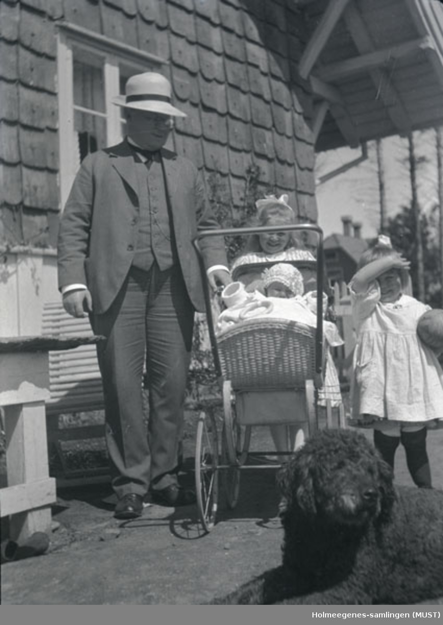 En mann og to små jenter pluss en i barnevogn foran huset på Holmeegenes i Stavanger. En svart hund ses i nedre høyre hjørne.