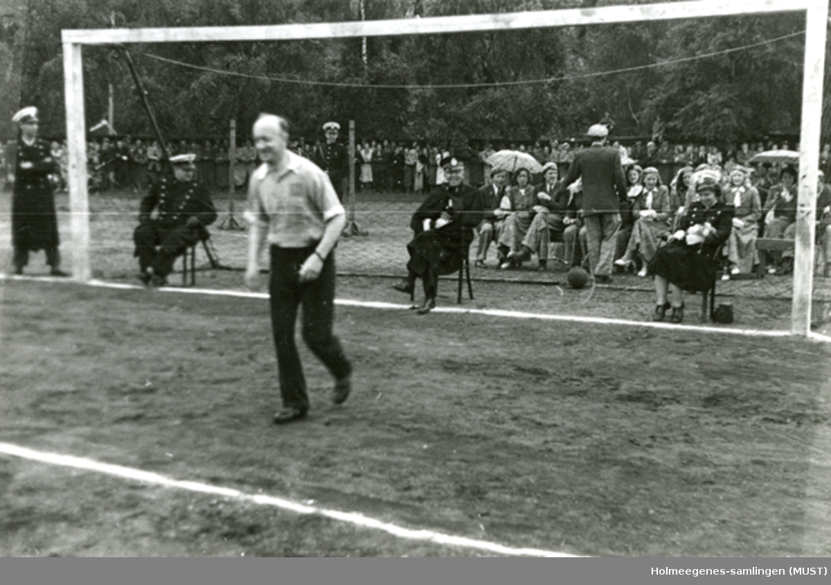 Tre politifolk sitter i et fotballmål. En mann springer foran målet. En gruppe mennesker sitter bak.
