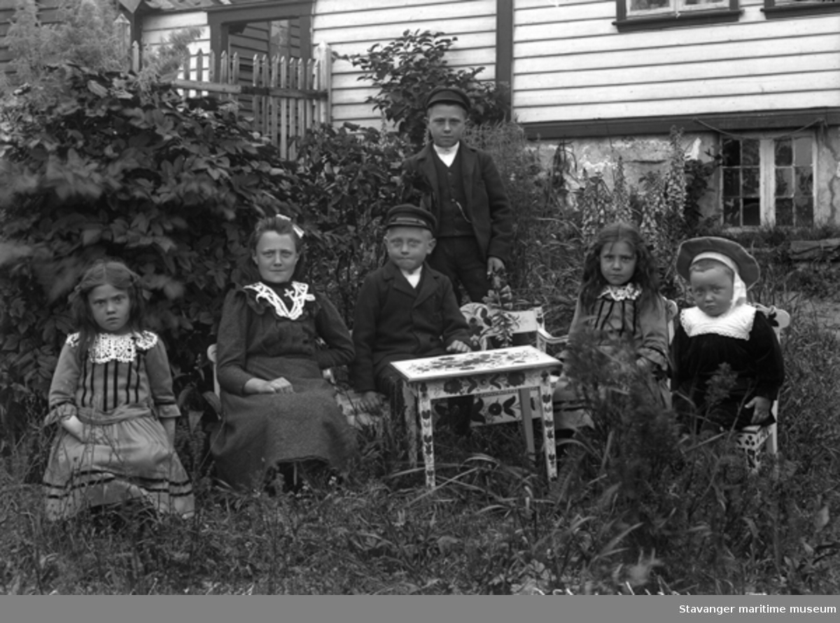 Gruppeportrett. Seks små barn sitter på barnemøbler i en frodig hage. Foto tatt på ettersommeren, ca 1901-1902. Platen merket Dusevig- Kirkhus' barn.