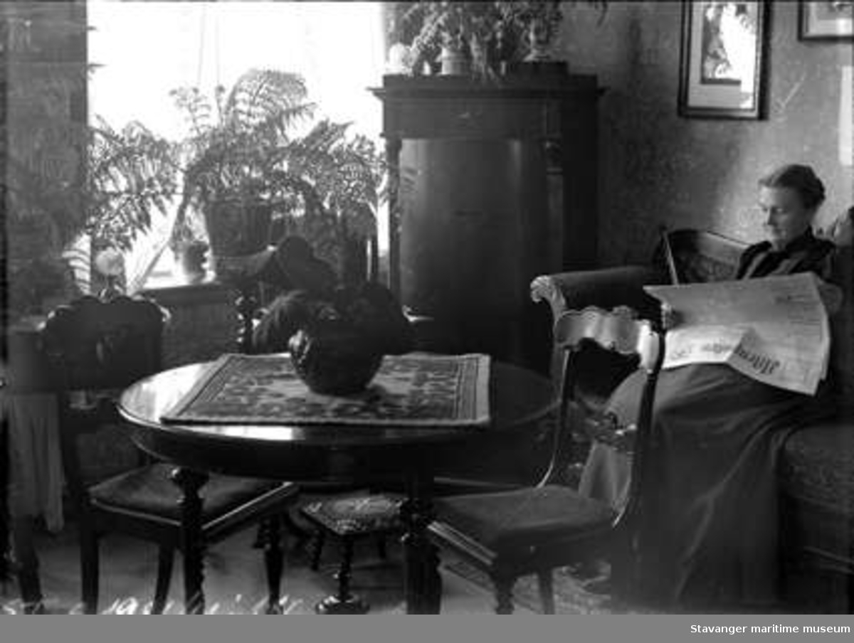Borgerlig interiør.Dame som sitter i sofa og leser Aftenposten. Rundt bord med tre stoler, pidestaller med bregnere.