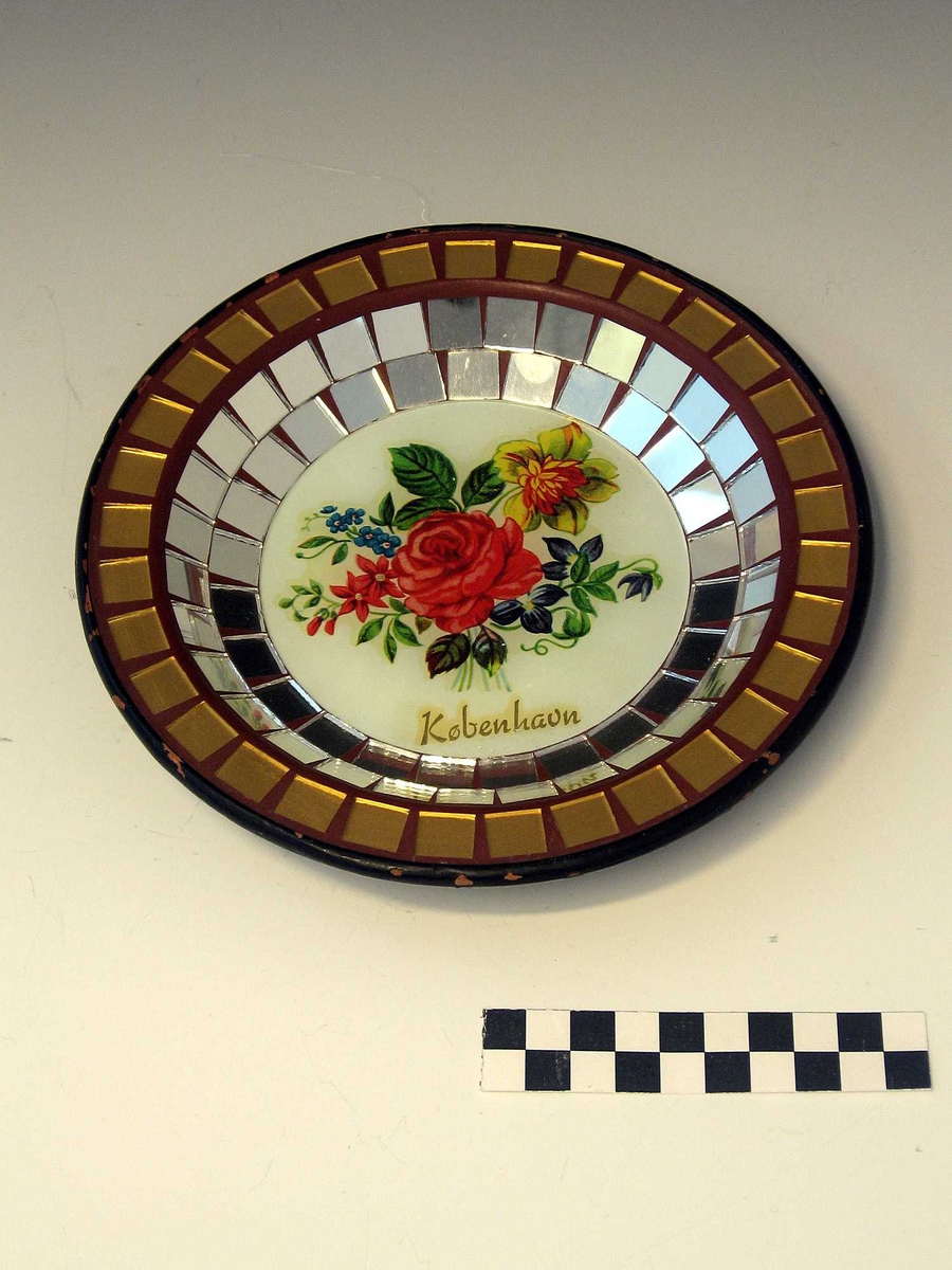 En typisk souvenir fra 1950-60-tallet. Innkjøpt i København. Selve tallerkenen er av keramikk, med mosaikk i glass og speil. Motivet i bunnen er dekket av glass.