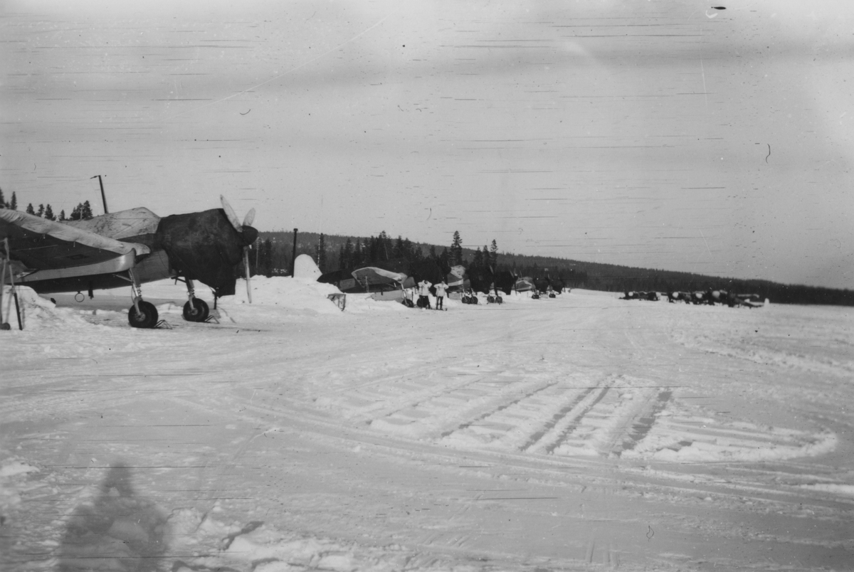Flera flygplan S 17 och S 18 på ett flygfält vintertid. Två män på skidor.