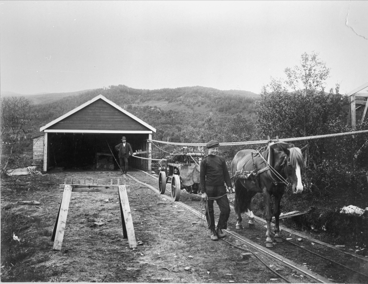 Gutt leier en hest med vogn på en skinnegang. Anton Hansen står i åpningen på reperbanebygningen til venstre.