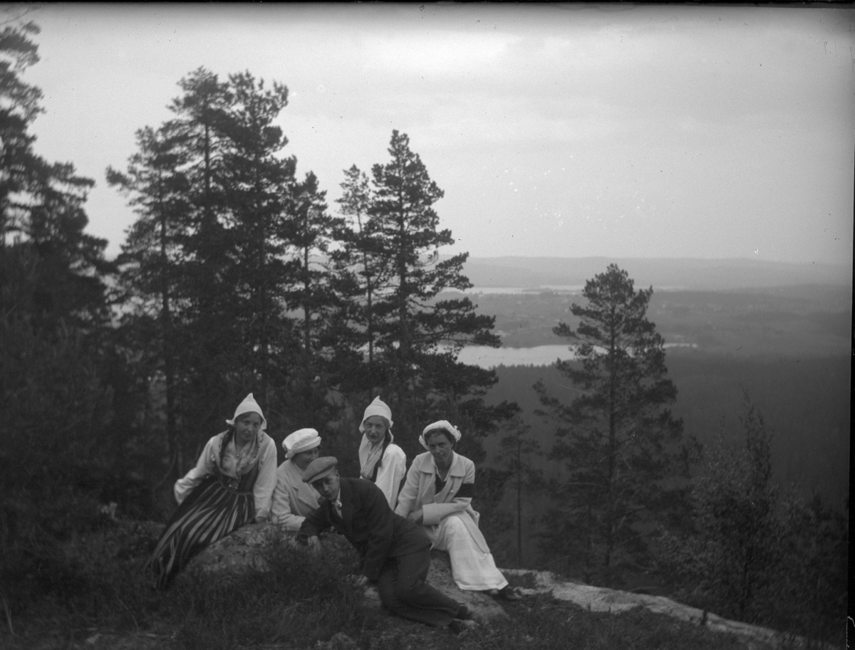 'Bildtext: ''Skorsberget.'' :: Gruppbild med 4 kvinnor och 1 man sittande vid berghäll på Skorsberget. Vy ned mot sjö, Insjön. Kvinnorna iklädda dräkter. Klädsel. ::  :: Ingår i serie med fotonr. 5223:1-13. Se även fotonr. 5231:1-11, 5232:1-8 och 5234:1-12.'