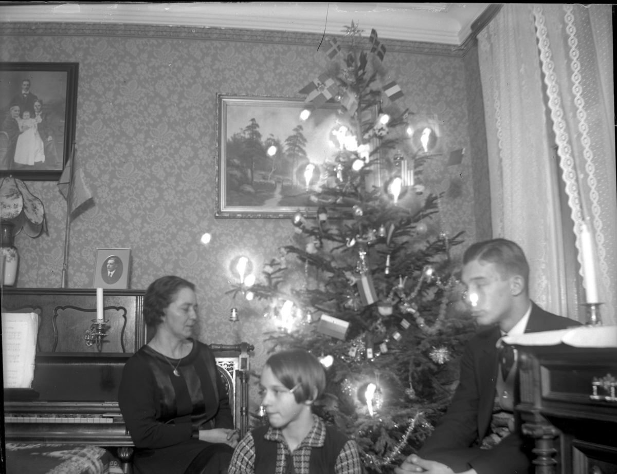 'Rum där 1 kvinna, 1 ung man och 1 flicka sitter vid en julgran med tända ljus och flaggor och glitter. I bakgrunden piano. I rummet tavlor, ljusstake, bordslampa, gardin, tapet. ::  :: Ingår i serie med fotonr. 5227:1-9.'