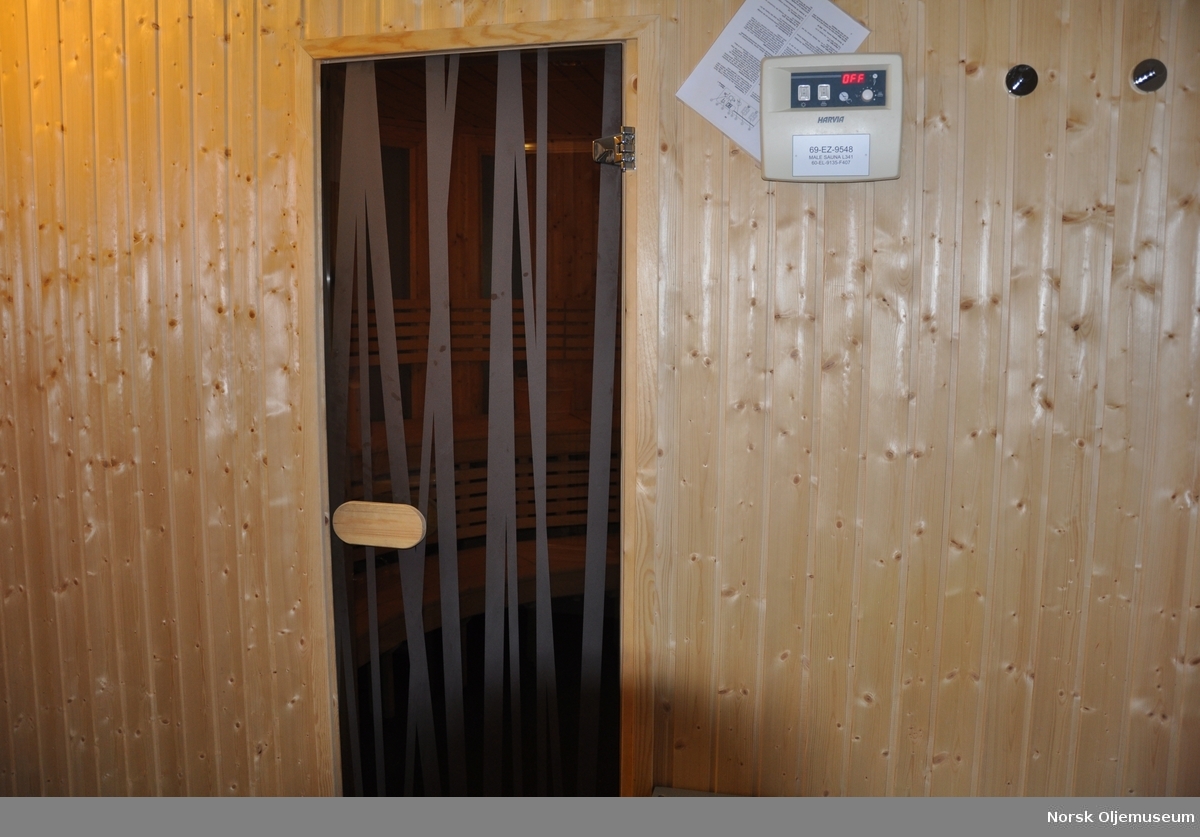 Dør inn til badstue på Valhallfeltet. Døra er dekorert med langsgående ujevne linjer. Til høyre for døra står et panel med bruksanvisning, antageligvis for styring av ovnen i badstua. Antageligvis prosess- og boligplattformen PH.