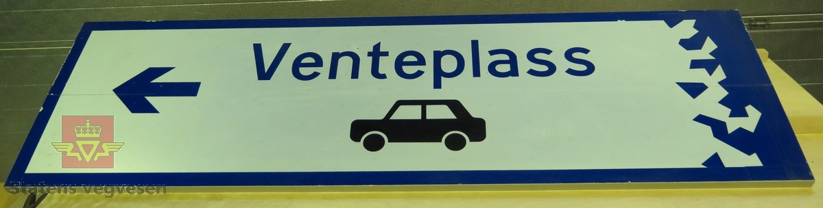 Rektangulært skilt av aluminium. Har symbol for kjøreretning og bil samt teksten "Venteplass". Flerfarget, med grå bakside.
