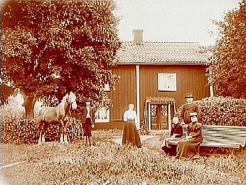 Tvåvånings bostadshus, närkesstuga. 
Familjegrupp 5 personer och en häst framför stugan.
C.O. Erikson.