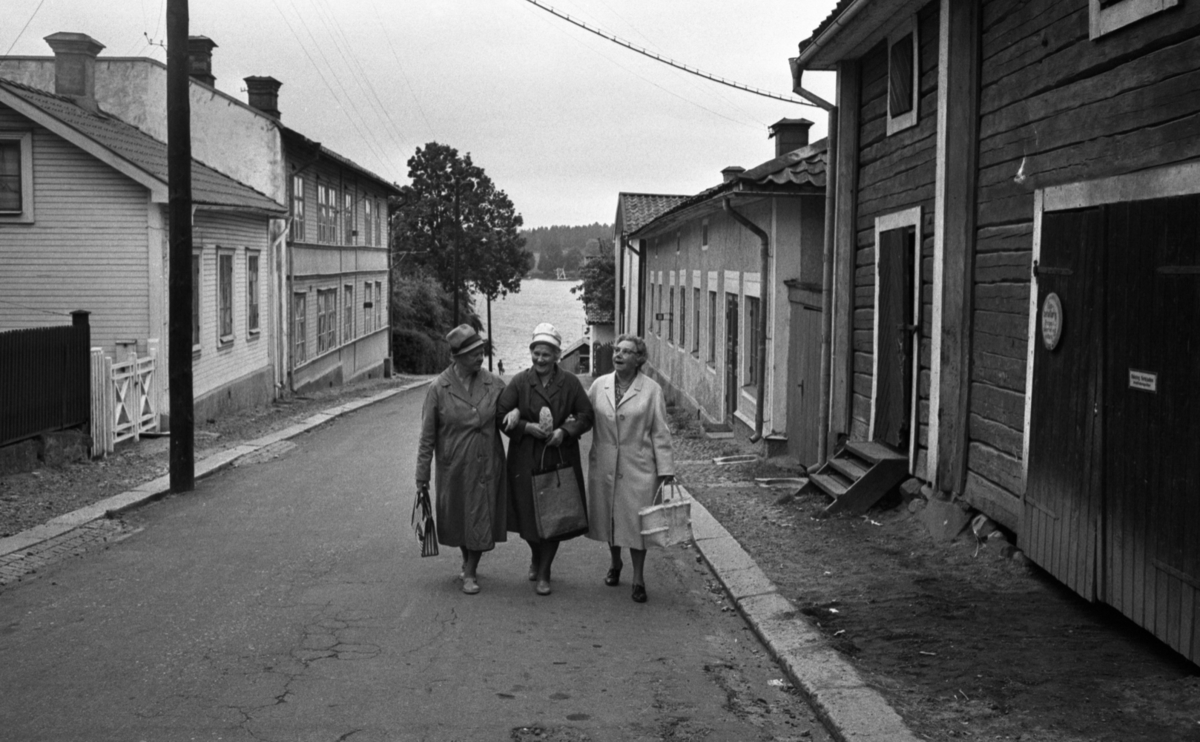 Noramarknad, Simmar i Gustavsvik 4 september 1965 

Tre äldre damer går arm i arm på en gata i Nora. De är klädda i kappor, två av dem bär hattar och alla bär handväskor i sina händer. Byggnader syns i bakgrunden.