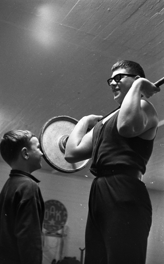 Tyngdlyftarreportage 31 januari 1966 

En pojke i övre tonåren klädd i sportlinne och sportbyxor samt med ett bälte i midjan lyfter en skivstång upp till axlarna inne i en träningslokal. Han sysslar med tyngdlyftning.  Han bär glasögon. Han ler mot en liten pojke i tioårsåldern som står och blickar upp mot honom.