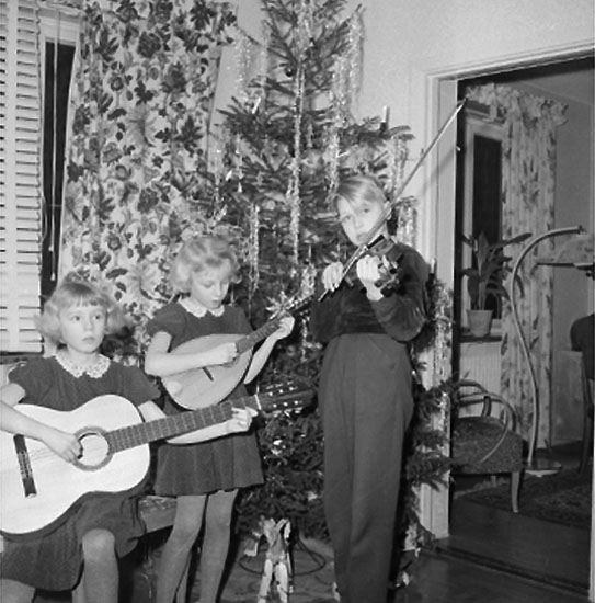 Rumsinteriör, grupp tre barn med musikinstrument vid julgranen.
Rulle Molin (beställare).