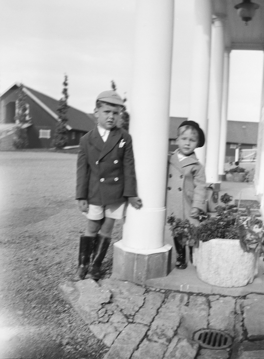 Pierre og Jan, på hver sin side av en søyle, ved inngangspartiet til hovedhuset på Linderud Gård. Begge har skitne gummistøvler.