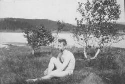 Naken mann sitter i gresset ved sjøen.