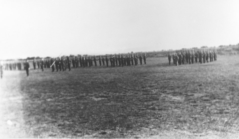 Vi antar at bildene er tatt i forbindelse med gjenopprettelse av Varanger batlajon 01.07.1934. Bildet viser oppstilte soldater på eksersersplassen på Nybergmoen 1934. Soldaterhjemmet i bakgrunnen.