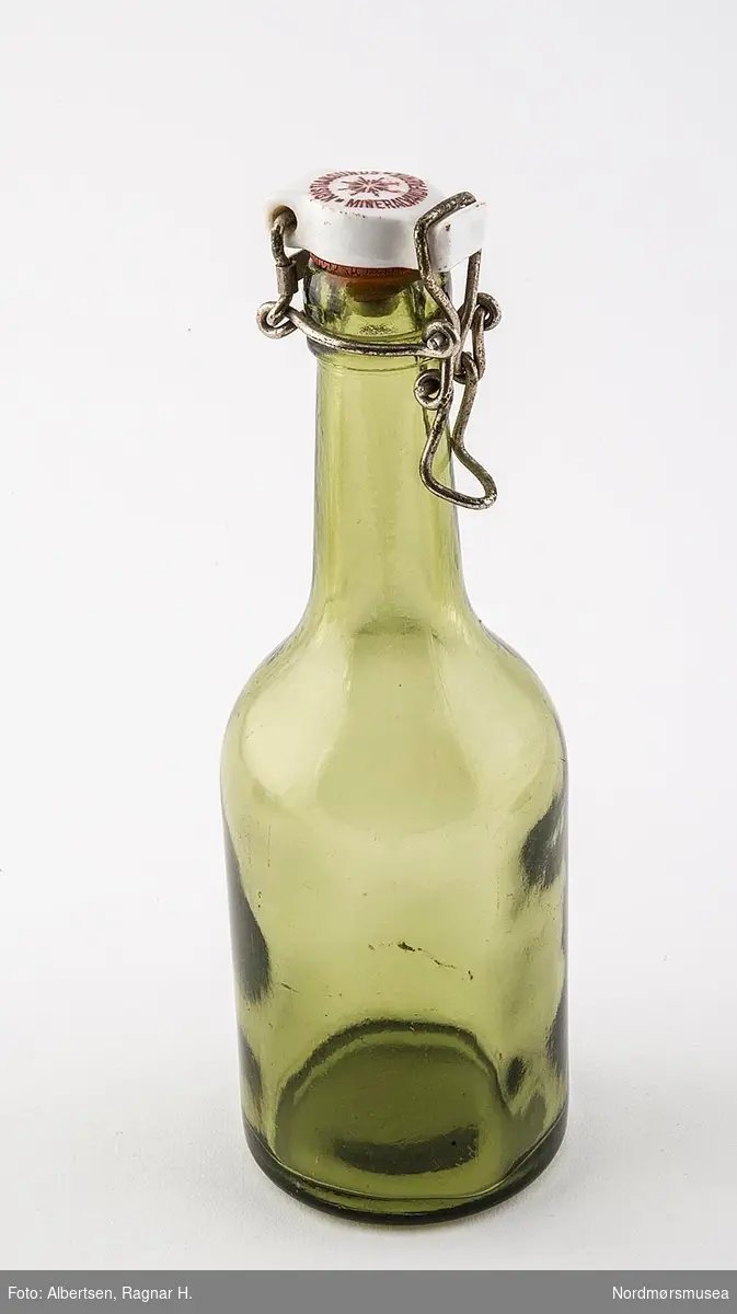 Rund grønn glassflaske med slake skuldre. Toppen er laget av steingods med korkdelen som stikkes ned i flasken. Toppa holdes med metall-hekting (svingtopp).