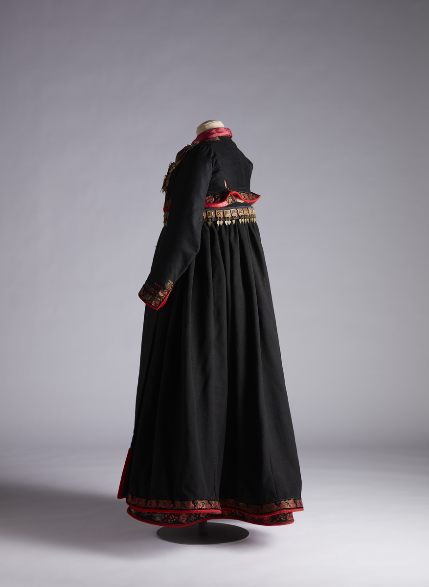 Forkle i svart ulldamask med kanting av rødt klede med sølvknipling og brosjert silkebånd med blomstermønster.