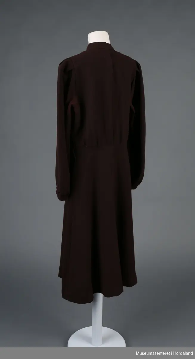 Form: langerma kjole med mansjettar, skrådd skjørt, sidelukking med trykk-knappar, og halslinning lukka med fire patentknappar bak  
