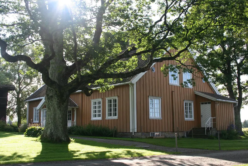 Bildet viser Gulbygningen fra siden. Tuntreet står litt i veien, men solen skinner gjennom og lyser opp.