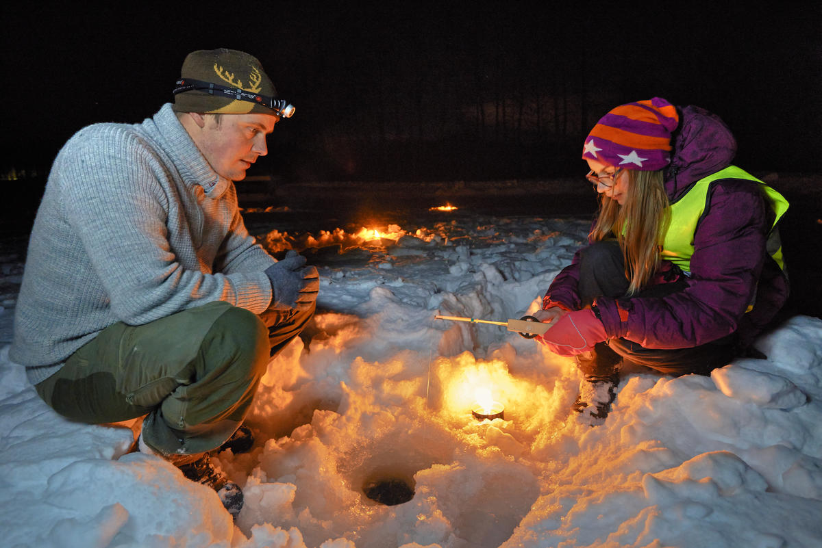 Bildet viser en mann og ei jente som sitter på isen å pilker. Det er mørkt og det har blitt tent opp fakkelbokser for å få lys.