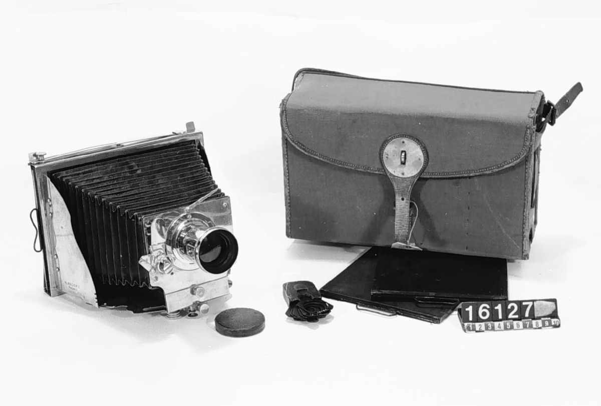 This was the first metal folding camera, the Model II. It was designed with a patent by H. Mader in 1888-1890. The camera was donated to the museum in January 1939 by Helmer Bäckström, who was a professor of photography. The camera is marked "5" in the slats of the bellows as well as with a round mark with the letters G M R and two sword-like characters. The lens is marked "Aplanal Invincible" and has "VI" engraved on the back at the threads. The camera comes with four cassettes, five plug-in apertures and a textile-clad case.