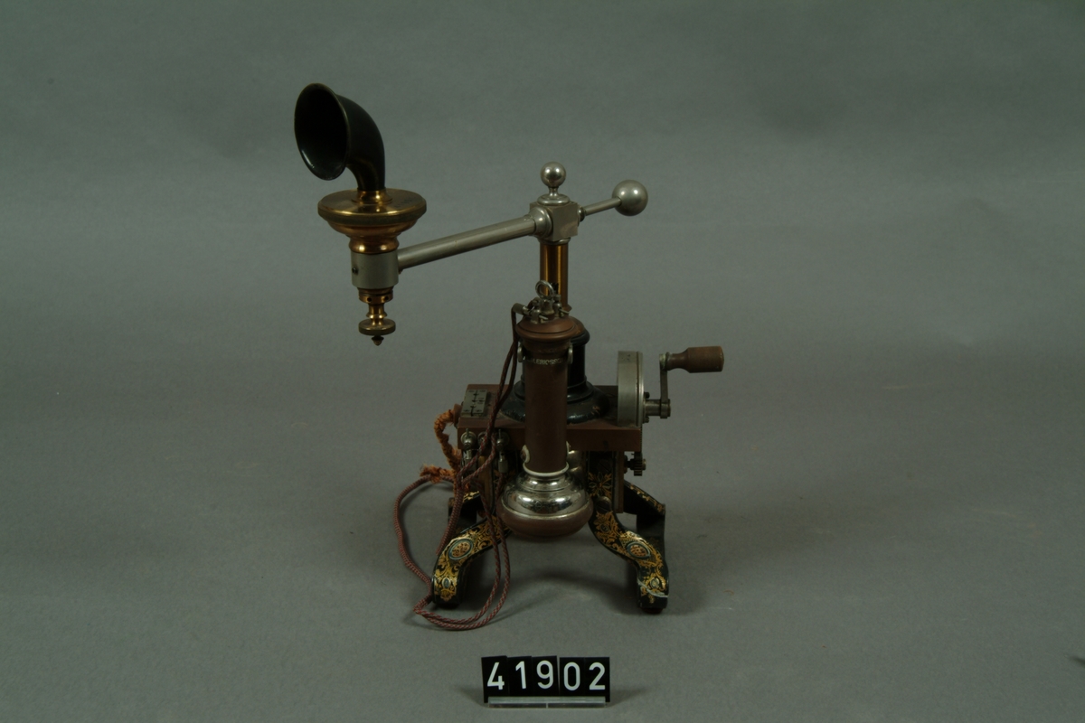 Telefonapparat, bordsmodell för lokalbatteri, med spiralmikrofon på svängbar arm. S.k. Tax. I denna telefon från 1884 utgörs de krumma benen av magneterna till ringsignalinduktorn. Den av Lars Magnus Ericsson konstruerade spiralmikrofonen sitter på en svängbar arm.