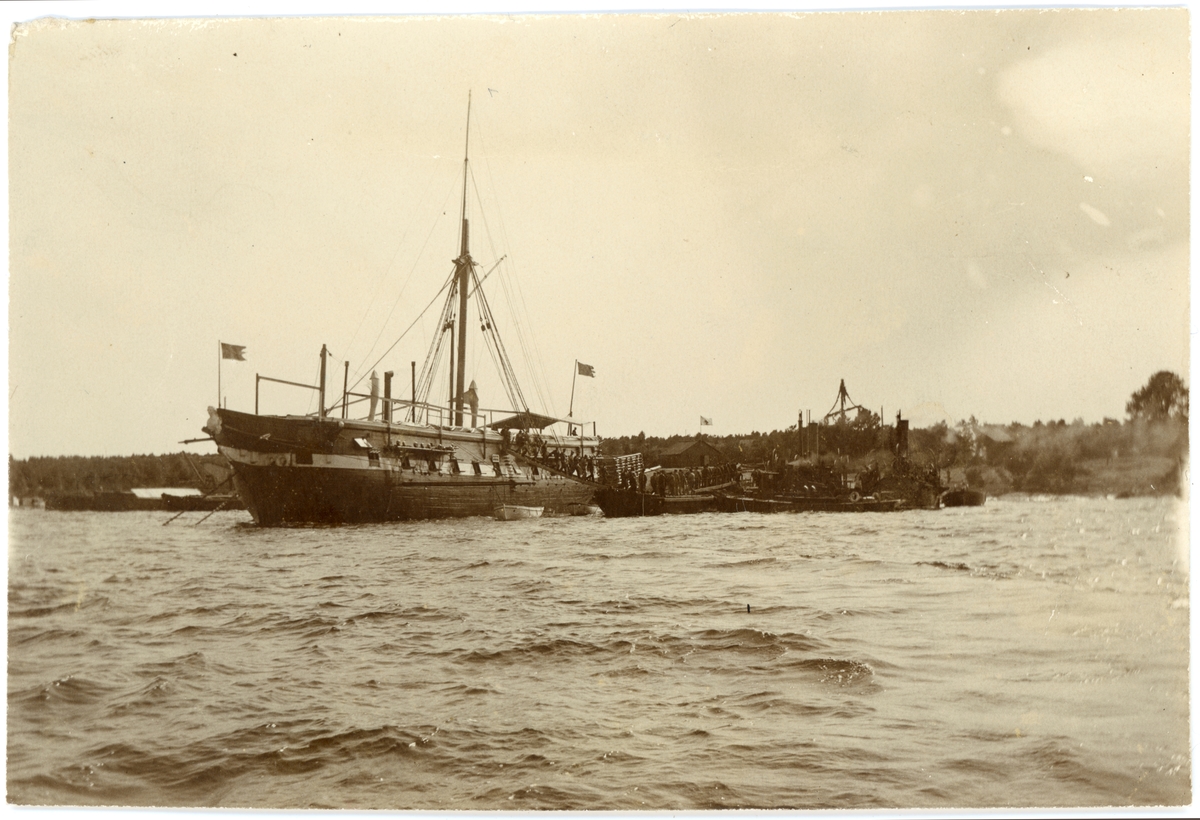 Bilden visar fregatten Af Chapman nedriggad efter ombyggnad till logementsfartyg. Vid sidan ligger flera små båtar förtöjd. En grupp soldater lämna fartyget genom landgången. I bakgrunden ser man en midsommarstång.